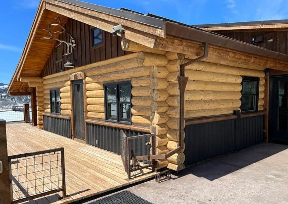 Log cabin exterior with wooden beams, deck, and large windows under a clear blue sky.