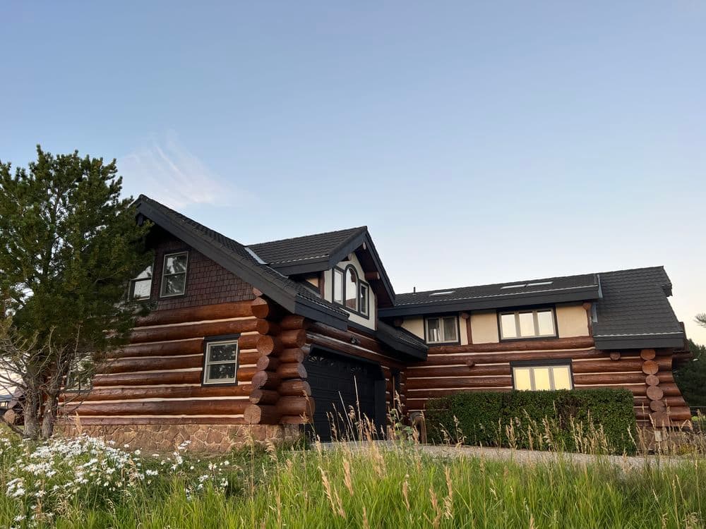 Log cabin home with a modern design, surrounded by greenery and flowers under a clear sky.