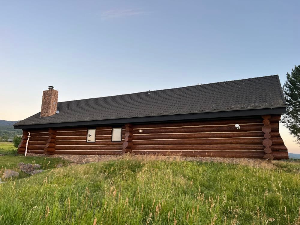 Log cabin house nestled in grassy landscape against a clear sky, featuring a stone chimney.