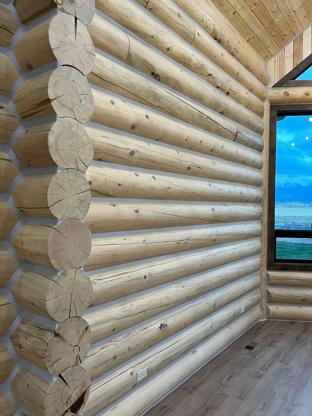 Interior view of a log cabin with natural wooden logs and a large window showcasing a scenic view.